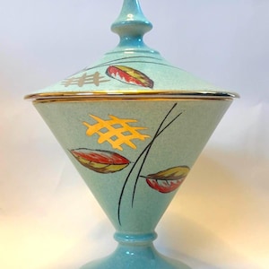 May include: A light blue conical lidded jar with a pedestal base. The surface features a speckled finish, gold-painted leaf motifs, and thin black line accents. The rim of the jar and the lid are trimmed with a thin gold band.