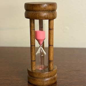 May include: A wooden hourglass with a clear glass center filled with pink sand. The hourglass is encased in a brown wooden frame with a rounded top and base. The sand is flowing from the top to the bottom.
