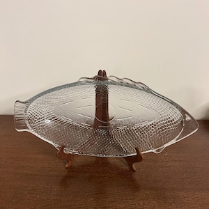 Peut inclure: Plat de service en verre transparent en forme de poisson, avec des écailles et des nageoires texturées. Le plat est présenté sur un support en bois. Un élément décoratif pour servir des apéritifs ou des collations.