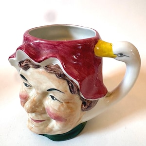 Può includere: Una tazza in ceramica a forma di personaggio con cuffia rossa, dotata di un manico a forma di collo d'oca bianco con becco giallo. Il volto presenta dettagli dipinti tra cui capelli castani, guance rosate e occhi scuri su base crema.