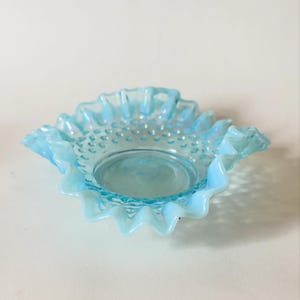 Puede incluir: Un cuenco de vidrio azul claro con un borde ondulado y un patrón de clavos. El cuenco tiene una base redonda y plana. El vidrio tiene un brillo iridiscente. Este cuenco decorativo es adecuado para guardar objetos pequeños.