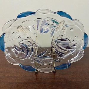 May include: A clear glass serving dish with a scalloped edge and blue trim. The dish features an embossed floral and seashell design. It is displayed on a decorative metal stand.