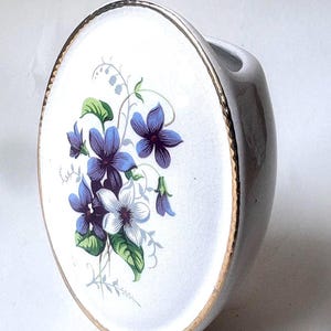 May include: An oval-shaped ceramic wall pocket vase with a white glazed finish and a fine gold-toned rim. The front surface features a detailed floral illustration of purple and white violets with green leaves and delicate trailing stems.