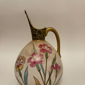 May include: An off-white ceramic pitcher with a gold handle and spout. The pitcher is decorated with pink, yellow, and red floral designs and green leaves. The spout has a black and gold patterned design.