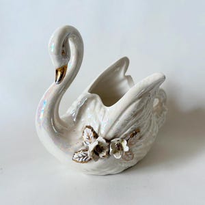Puede incluir: Una figura de cisne de cerámica blanca con detalles iridiscentes. El cisne tiene un cuello curvado y alas abiertas, con detalles dorados en el pico. Flores decorativas con detalles dorados adornan el lateral. Una pieza decorativa.