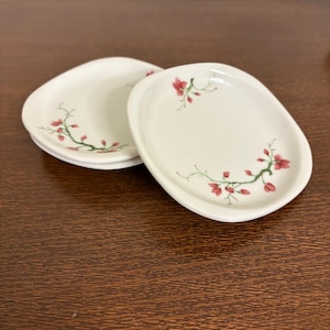 Puede incluir: Un juego de cuatro platos pequeños de cerámica blanquecina. Cada plato presenta un diseño floral con flores rojas y tallos verdes. Los platos tienen una forma irregular y están apilados sobre una superficie de madera oscura.