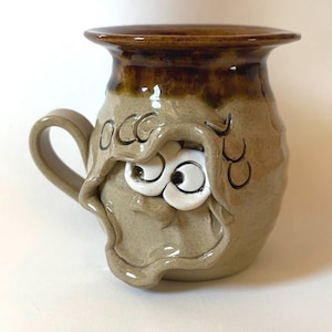 May include: A tan ceramic mug with a dark brown glazed rim and a handle on the left side. The mug features a sculpted face with two large white eyes peeking out from under a draped, hood-like clay fold. The letters OCC are etched into the clay.