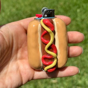 May include: A novelty lighter case shaped like a hot dog in a bun, featuring a brown sausage topped with wavy lines of red ketchup and yellow mustard. The lighter mechanism is visible at the top, with a small red ignition button and metal casing.
