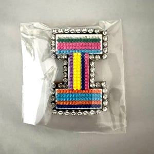 May include: A colorful brooch in a clear plastic bag. The brooch has a rectangular top and bottom connected by a narrower vertical section. It features a rainbow of colors and is outlined with clear rhinestones.