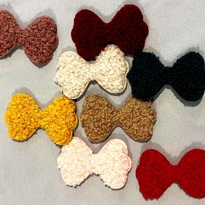 May include: A collection of eight textured bow hair clips in various colors. The bows are in shades of brown, red, white, black, yellow, and pink. The bows are made of a soft, fuzzy material.