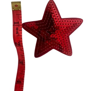 May include: A red sequined star with a 10 cm measurement next to it. The star is a vibrant red colour and is covered in small, shiny sequins. The measuring tape is red with white markings.