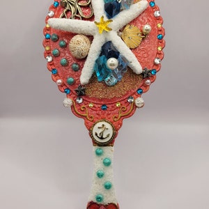 May include: A red hand mirror with a white starfish, seashells, and blue and white glass beads. The handle is white with blue dots and a red heart with a gold anchor.