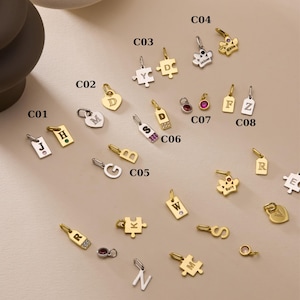 May include: A selection of gold and silver charms, including letter and shape pendants. Some charms feature gemstones or engraved names. The charms are displayed on a neutral surface, showcasing various designs.