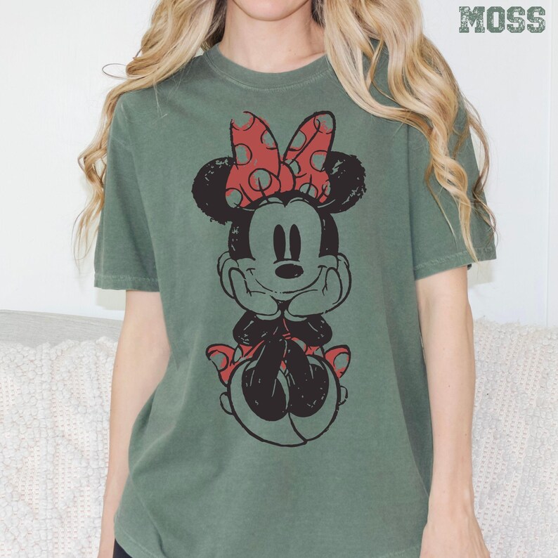 May include: Green t-shirt featuring a distressed graphic of Minnie Mouse with a red polka dot bow. The word "MOSS" is printed in green at the top right. The shirt has short sleeves and a crew neck.