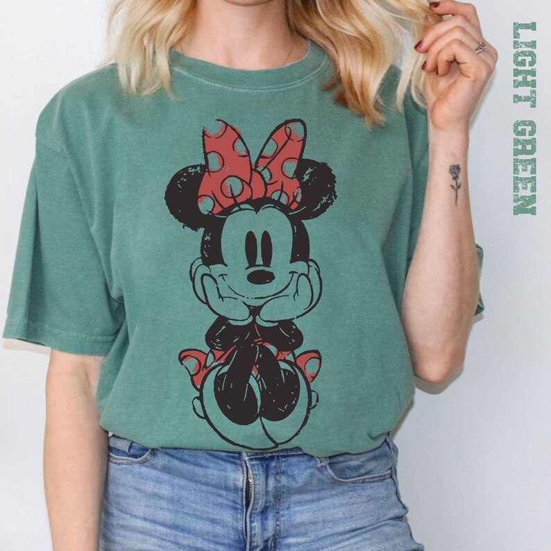 May include: Light green t-shirt featuring a distressed graphic of Minnie Mouse. Minnie is depicted in black with a red polka dot bow and accents. The shirt has a relaxed fit and short sleeves, perfect for casual wear.