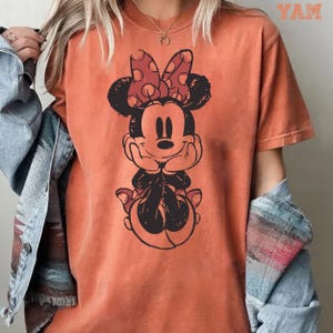 May include: Orange t-shirt featuring a distressed graphic of Minnie Mouse with a red polka dot bow. The cartoon character is smiling and resting her chin on her hands. The word "YAM" is printed in the upper right corner.