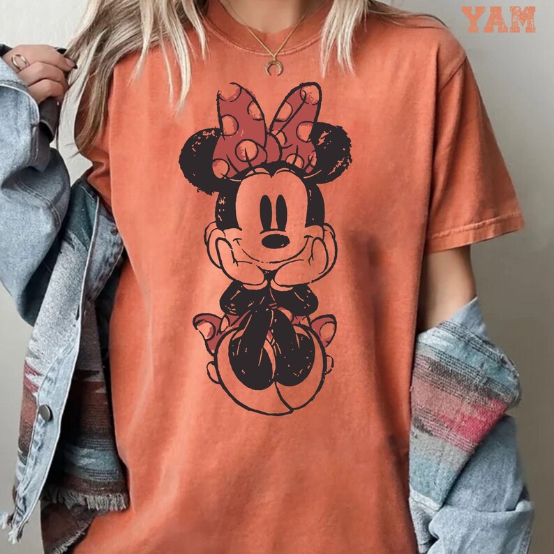 May include: Orange t-shirt featuring a distressed graphic of Minnie Mouse with a red polka dot bow. The cartoon character is smiling and resting her chin on her hands. The word "YAM" is printed in the upper right corner.