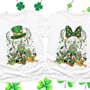 May include: Two white t-shirts featuring a St. Patrick's Day design. One shirt has a green leprechaun hat and the other a green bow, both with Mickey Mouse ears. The shirts also feature Disney characters and a castle.