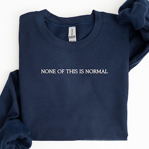 May include: Navy blue sweatshirt with the phrase "NONE OF THIS IS NORMAL" printed in white. The sweatshirt is made of a soft material and has a crew neck design. The Gildan brand tag is visible.