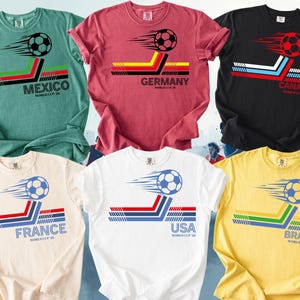 May include: A collection of six t-shirts in various colors, each featuring a soccer ball graphic with country flags and text. The shirts include designs for Mexico, Germany, Canada, France, USA, and Brazil, with the text "WORLD CUP 20" below.