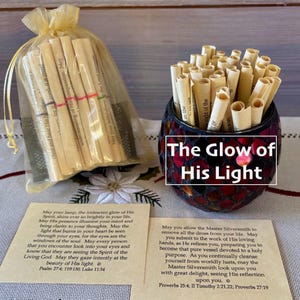 May include: A collection of rolled paper scrolls, some in a clear bag tied with a ribbon, and others in a decorative glass jar. The text "The Glow of His Light" is displayed on the jar. Two cards with text are also visible.