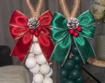 May include: Two hanging ornaments featuring clusters of wooden spheres, one white and one dark green. Each ornament has a jute rope loop, a large satin bow with glittery edges, a small pinecone, and three red berries at the top center.