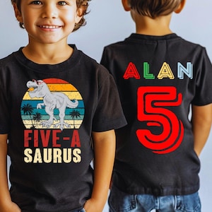 May include: Black t-shirt with a dinosaur graphic and the text "FIVE-A-SAURUS" in red and white. The back of the shirt has the name "ALAN" in colorful letters and the number "5" in red. The shirt is worn by a young child.