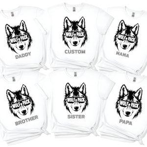 May include: White t-shirts with a black wolf graphic wearing sunglasses that say "WOLF PACK." Each shirt has a different word below the wolf: DADDY, CUSTOM, NANA, BROTHER, SISTER, and PAPA. The shirts are short-sleeved.