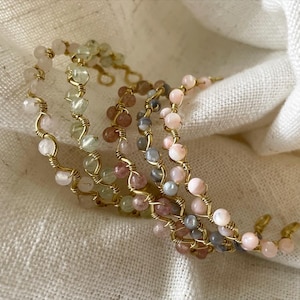 May include: Close-up of several beaded bracelets. Each bracelet features small, colorful beads in shades of pink, green, and gray, strung on gold-colored wire. The bracelets are arranged on a white, textured surface.
