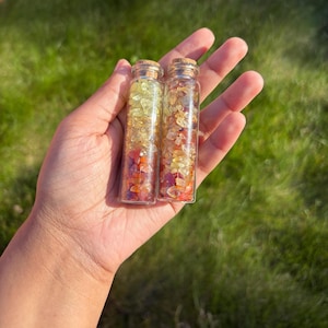 May include: Two small glass bottles with cork stoppers, filled with layers of colorful gemstones. The gemstones include shades of yellow, orange, and red. The bottles are held in a hand against a blurred green background.