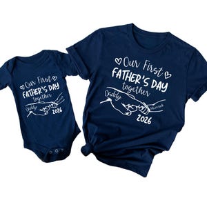May include: Navy blue baby onesie and t-shirt set. Both garments feature the text "Our First Father's Day together" with a graphic of two hands holding each other. The text "Daddy" and "Maverick 2026" are also included.