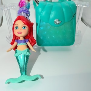 May include: Ariel-themed keychain featuring a teal backpack and a doll of Ariel with red hair, a purple top, and a teal mermaid tail. The keychain includes a beaded charm with pink, purple, and blue beads. The backpack has a seashell-shaped clasp.