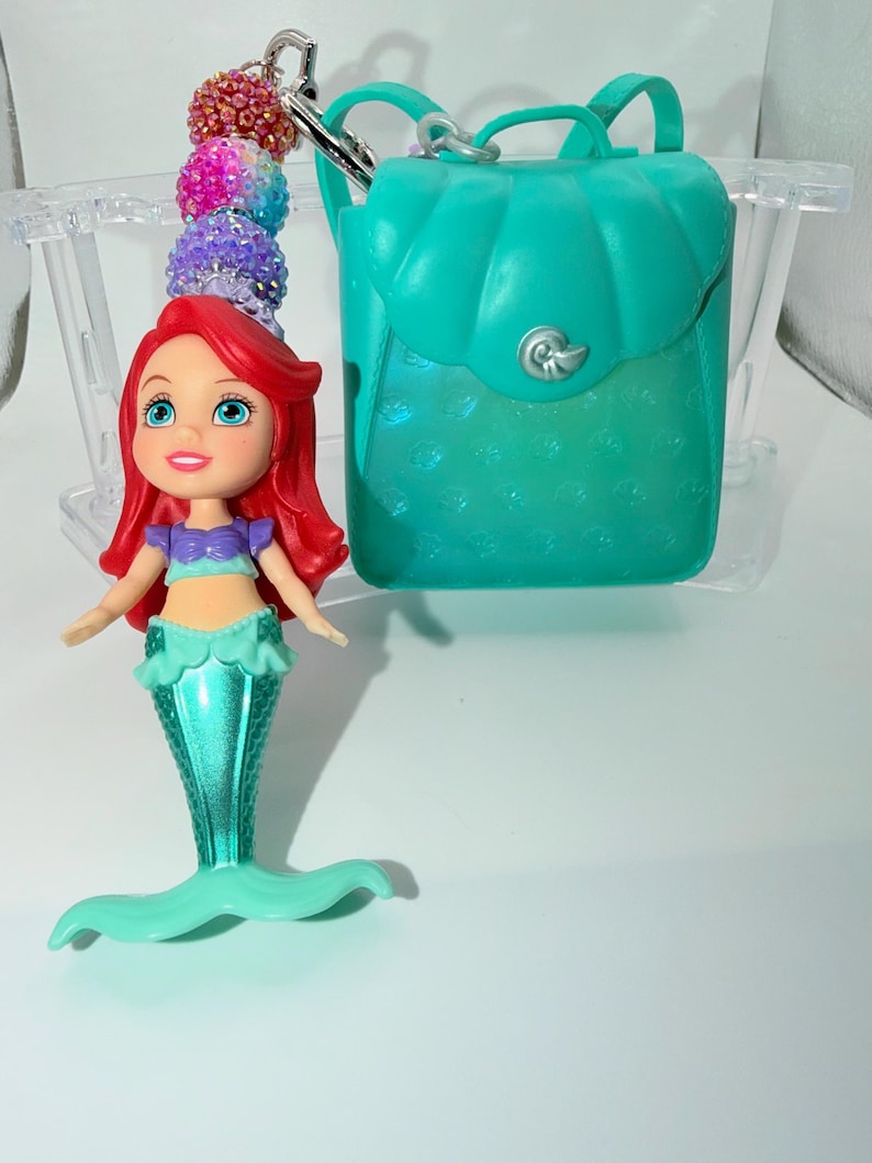 May include: Ariel-themed keychain featuring a teal backpack and a doll of Ariel with red hair, a purple top, and a teal mermaid tail. The keychain includes a beaded charm with pink, purple, and blue beads. The backpack has a seashell-shaped clasp.
