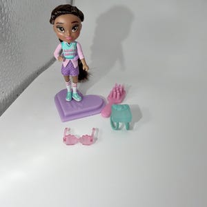 May include: A doll with brown hair and a pink and teal outfit stands on a purple heart-shaped base. Accessories include a pink hairbrush, a teal backpack, and pink sunglasses. The doll's shirt reads "SHINGLE HOPPER".