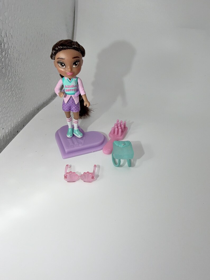 May include: A doll with brown hair and a pink and teal outfit stands on a purple heart-shaped base. Accessories include a pink hairbrush, a teal backpack, and pink sunglasses. The doll's shirt reads "SHINGLE HOPPER".