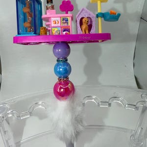 May include: A decorative pen with a miniature pet shop scene, including a pink and purple platform, a blue and orange aquarium, and a dog house. The pen has colourful beads and a fluffy white tail.