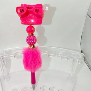 May include: A vibrant pink pen featuring a View-Master design, embellished with pink beads and a fluffy pink pom-pom. The pen rests in a clear acrylic holder.