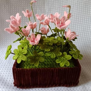 Può includere: Composizione floreale decorativa con fiori rosa e fogliame verde in un contenitore rettangolare marrone. I fiori hanno petali delicati e le foglie sono di un verde vibrante. Il contenitore è realizzato in un materiale marrone scuro strutturato.
