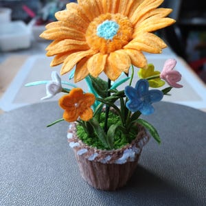 Op de afbeelding: Een handgemaakt vilten bloemstuk in een kleine pot. Het arrangement heeft een grote oranje zonnebloem met een blauw centrum, omringd door kleinere kleurrijke bloemen in een bruine pot met witte rand. Groene bladeren en stelen maken het ontwerp compleet.