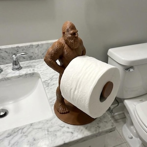 May include: A brown, sculpted figure of a hairy, bipedal creature resembling a Bigfoot or Sasquatch, standing on a circular base. The figure holds a horizontal rod that supports a single roll of white toilet paper with a floral pattern.