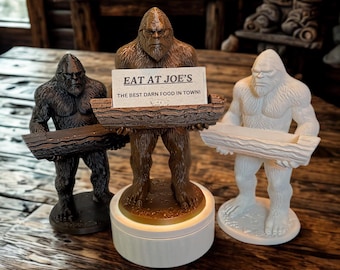 May include: Three Bigfoot statues in varying colors: dark brown, brown, and white. The brown statue holds a sign that reads "EAT AT JOE'S THE BEST DARN FOOD IN TOWN!" Each statue is holding a wooden log.