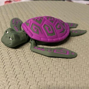 Puede incluir: Una figura de tortuga impresa en 3D en tonos verdes y morados. El caparazón de la tortuga es de un morado vibrante con un diseño geométrico, mientras que la cabeza, las aletas y el cuerpo son verdes. La figura descansa sobre una superficie texturizada.