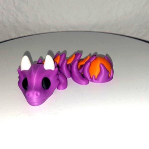 Könnte beinhalten: Ein lila, bewegliches Drachenspielzeug mit orangefarbenen und weißen Akzenten. Der Drache hat ein detailliertes, strukturiertes Design mit großen schwarzen Augen und weißen Hörnern. Der Körper des Drachen ist segmentiert, was eine flexible Bewegung ermöglicht.