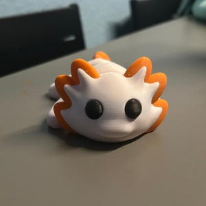 Könnte beinhalten: Eine 3D-gedruckte Figur, die ein weißes Wesen mit orangefarbenen Akzenten und großen schwarzen Augen darstellt. Das stilisierte Design hat eine stachelige Kontur. Die Figur liegt auf einer grauen Oberfläche. Die orangefarbenen Details heben Kopf und Rücken hervor.