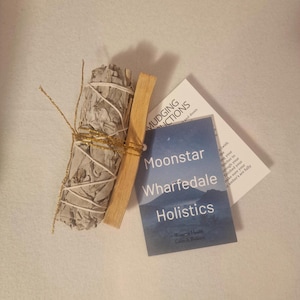 May include: A bundle of white sage wrapped with white string and gold twine, next to a piece of palo santo wood and a card with the text "Moonstar Wharfedale Holistics". The items are on a white surface.