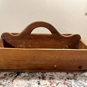 May include: A rectangular wooden tote with a curved handle. The open-top box is made of light brown wood with visible grain and some white paint flecks. The handle is arched, and the box has a simple, rustic design.
