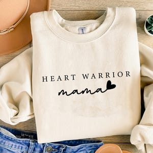 May include: A sand-colored sweatshirt with the text "HEART WARRIOR mama" in black. The sweatshirt is folded, with a pair of blue jeans and a brown hat in the background. A small succulent plant is also visible.