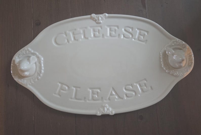 May include: Off-white oval cheese serving platter with embossed text reading "CHEESE PLEASE". The platter features decorative mouse head accents on each end. A charming addition to any kitchen.