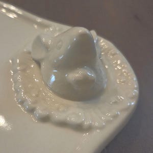 May include: A white ceramic serving dish with a raised, detailed mouse head on one end. The mouse head is smooth and glossy, surrounded by a decorative, embossed border. The dish has a curved, organic shape.