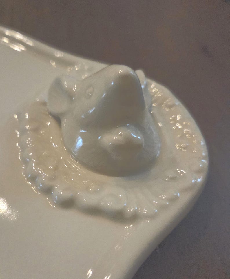 May include: A white ceramic serving dish with a raised, detailed mouse head on one end. The mouse head is smooth and glossy, surrounded by a decorative, embossed border. The dish has a curved, organic shape.